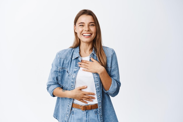 Bella giovane donna che ride e si diverte, ridacchia mentre si tiene per mano sul petto e sorride denti bianchi, esprime felicità ed emozioni positive, bianco