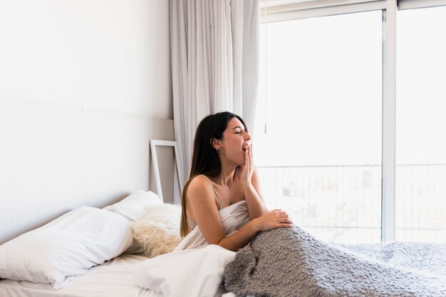 Bella giovane donna che si siede sul letto che sbadiglia