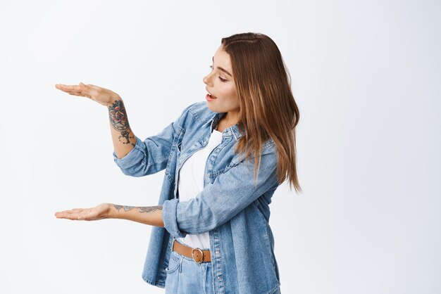 Bella giovane donna che tiene l'oggetto nelle mani che modellano una scatola di grandi dimensioni e guardandolo eccitato in piedi in abiti casual su sfondo bianco