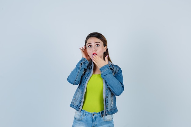 Bella giovane donna che tiene la mano sulla bocca mentre ascolta il segreto in abito di jeans e sembra sorpresa, vista frontale.