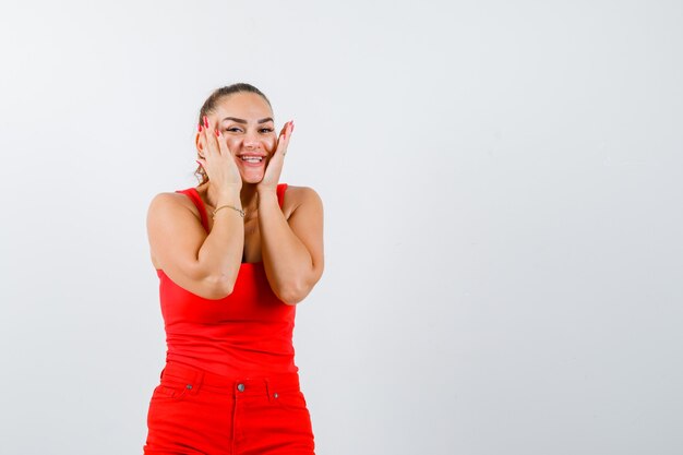 Bella giovane donna che tiene le mani sulle guance in canottiera rossa, pantaloni e che sembra allegra. vista frontale.