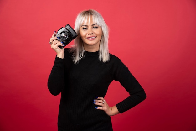 Bella giovane donna che tiene una macchina fotografica mentre levandosi in piedi contro il fondo rosso. Foto di alta qualità