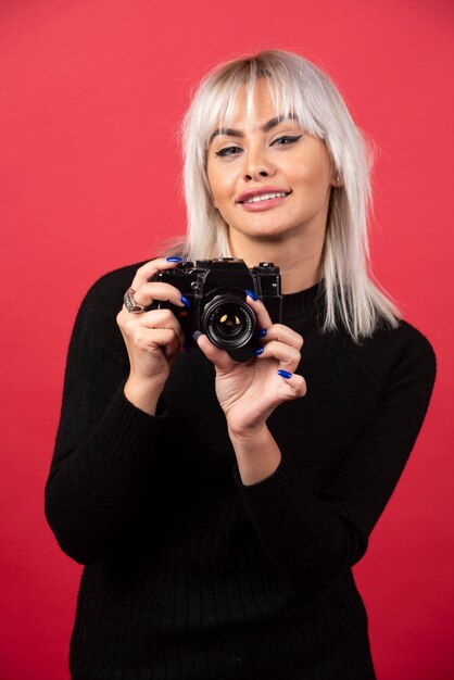 Bella giovane donna che tiene una macchina fotografica mentre levandosi in piedi contro il fondo rosso. Foto di alta qualità