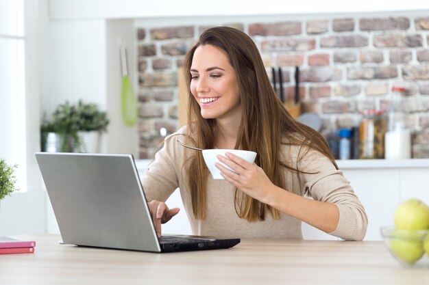 Bella giovane donna che utilizza il suo computer portatile in cucina.