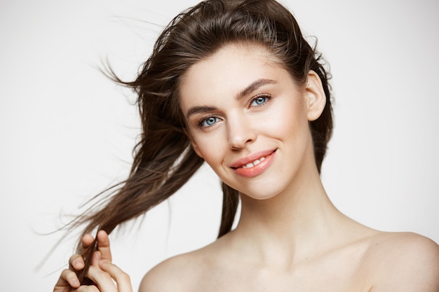 Bella giovane donna con i capelli commoventi sorridenti della pelle pulita perfetta sopra la parete bianca. Trattamento facciale.