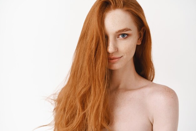 Bella giovane donna con lunghi capelli rossi perfetti e occhi azzurri che guardano davanti, in piedi nuda, che mostra la pelle pulita pallida e un taglio di capelli naturale, muro bianco