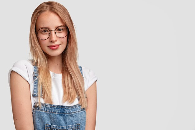 Bella giovane donna con un'espressione del viso soddisfatta, ha i capelli lunghi, la pelle sana, indossa occhiali rotondi e salopette di jeans