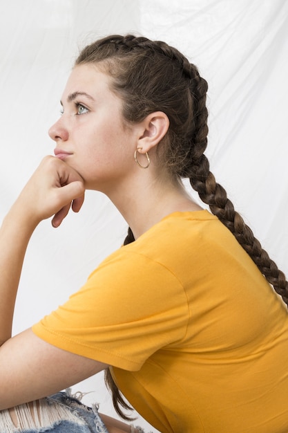 Bella giovane donna con una camicia gialla e capelli intrecciati seduta contro un muro bianco