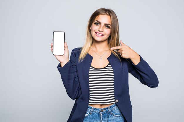 Bella giovane donna felice che tiene telefono cellulare dello schermo in bianco e dito puntato sopra il muro bianco