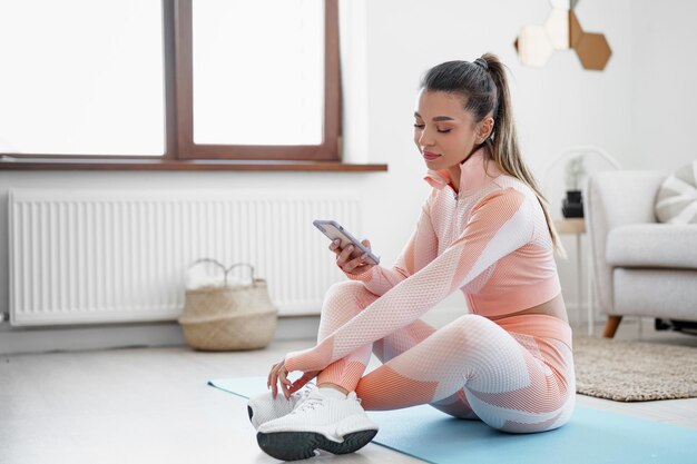 Bella giovane donna in abbigliamento sportivo seduta in casa e guardando lo smartphone prima dell'allenamento