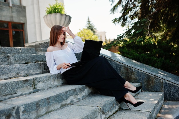 Bella giovane donna in camicetta bianca ampi pantaloni neri e tacchi alti classici neri seduta sulle scale e lavorando sul suo laptop