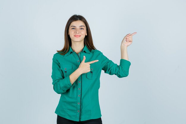 Bella giovane donna in camicia verde che punta a destra e sembra allegra, vista frontale.