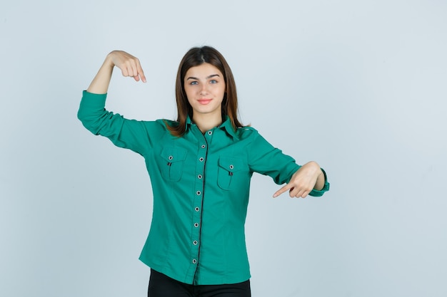 Bella giovane donna in camicia verde che punta a se stessa e guardando fiducioso, vista frontale.