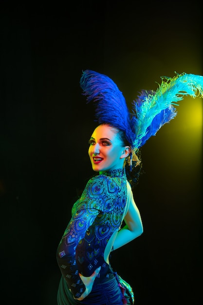 Bella giovane donna in carnevale, elegante costume in maschera con piume sul muro nero in luce al neon