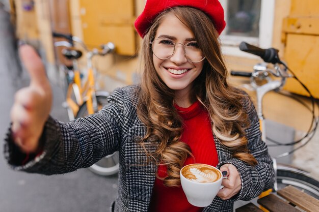 Bella giovane donna in giacca grigia che beve caffè con piacere in un caffè all'aperto