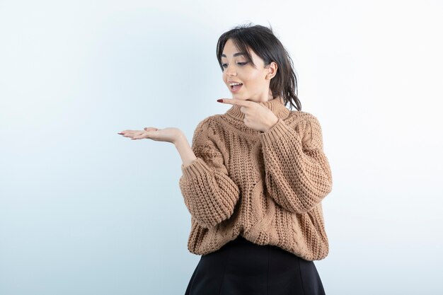 Bella giovane donna in maglione lavorato a maglia in piedi e in posa sul muro bianco