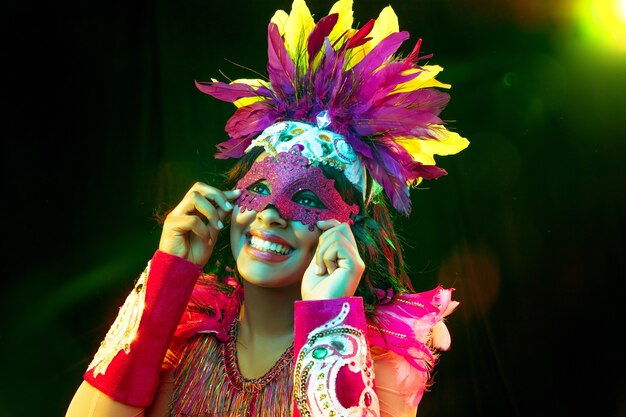 Bella giovane donna in maschera di carnevale ed elegante costume in maschera con piume in luci colorate e bagliore su sfondo nero