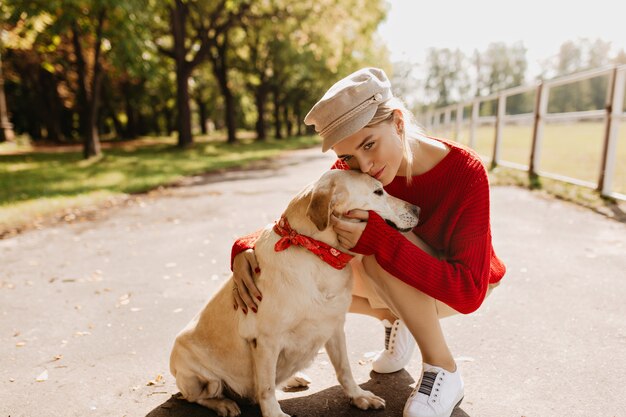 Bella giovane donna in vestiti alla moda che bacia teneramente il suo cane. Bella bionda con il suo animale domestico che gode del tempo soleggiato nel parco.