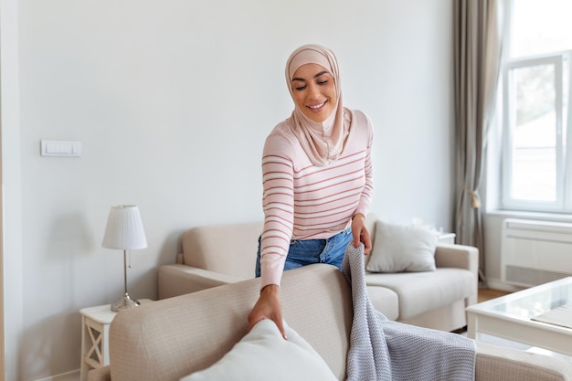 Bella giovane donna musulmana araba che mette morbidi cuscini e plaid sul comodo divano rendendo la sua casa accogliente e calda