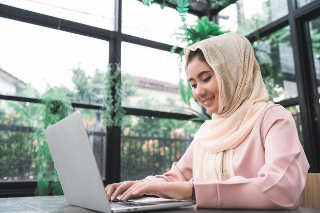 Bella giovane donna musulmana asiatica sorridente che lavora al computer portatile che si siede nel salone a casa