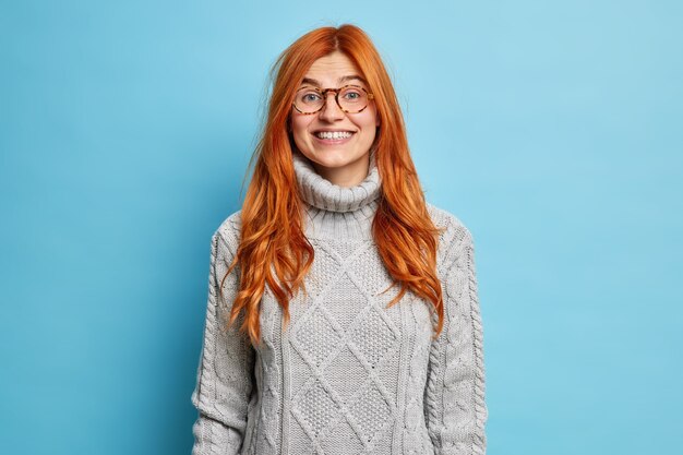 Bella giovane donna rossa con un aspetto europeo sorride piacevolmente vestita con un maglione caldo esprime emozioni positive e sente buone notizie.