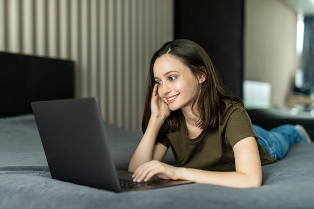 Bella giovane donna sdraiata sul letto e utilizzando il computer portatile per l'utilizzo dei social media e lo shopping online rilassarsi nell'accogliente camera da letto in vacanza