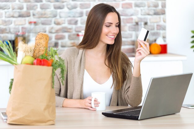 Bella giovane donna shopping con la sua carta online in cucina.