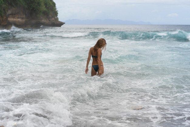 Bella giovane donna snella con lunghi capelli biondi in un costume da bagno sulla spiaggia vicino all'oceano. Rilassatevi sulla spiaggia. Vacanze tropicali. Una donna entra in acqua per nuotare.