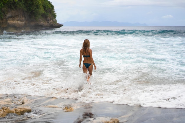 Bella giovane donna snella con lunghi capelli biondi in un costume da bagno sulla spiaggia vicino all'oceano. Rilassatevi sulla spiaggia. Vacanze tropicali. Una donna entra in acqua per nuotare.