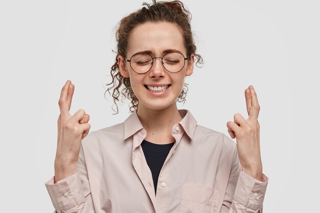 Bella giovane donna sognante e speranzosa tiene le dita incrociate, gli occhi chiusi, esprime desiderio sotto la stella cadente, indossa una camicia oversize beige e occhiali, isolato sopra il muro bianco