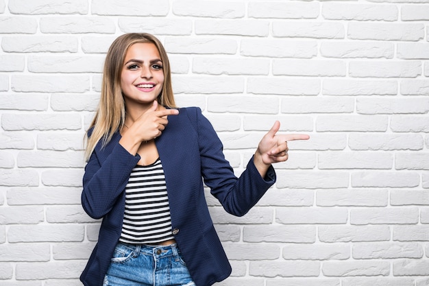 Bella giovane donna sopra il muro di mattoni bianco che sorride con la faccia felice che osserva e che indica al lato