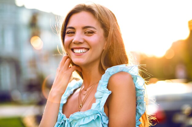 Bella giovane donna sorridente beata che posa in strada, luce solare intensa, vestito blu elegante alla moda, trucco naturale e umore positivo.