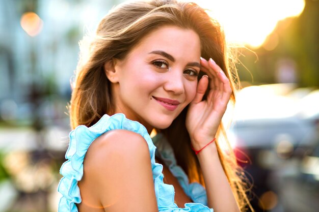 Bella giovane donna sorridente beata che posa in strada, luce solare intensa, vestito blu elegante alla moda, trucco naturale e umore positivo.