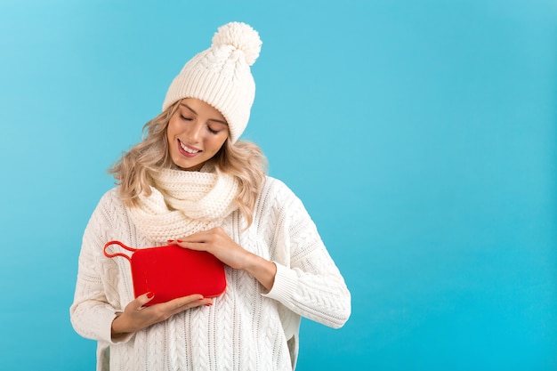 Bella giovane donna sorridente bionda alla moda che tiene altoparlante senza fili che ascolta la musica maglione bianco da portare felice e cappello lavorato a maglia che posa sull'azzurro