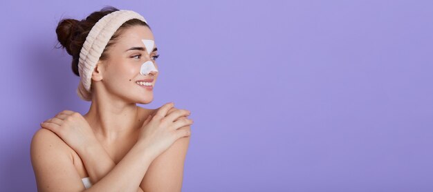 Bella giovane donna sorridente con la pelle perfetta pulita che guarda sognante da parte e tocca la sua spalla nuda, facendo procedure di bellezza