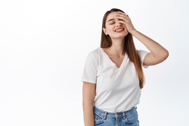 Bella giovane donna spensierata che tocca la pelle pulita idratata e perfetta, sorride e chiude gli occhi con espressione romantica, sogna ad occhi aperti, in piedi su sfondo bianco