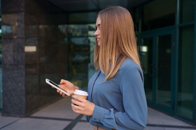 Bella giovane donna sta utilizzando un'app nel suo dispositivo smartphone per inviare un messaggio di testo vicino a edifici commerciali