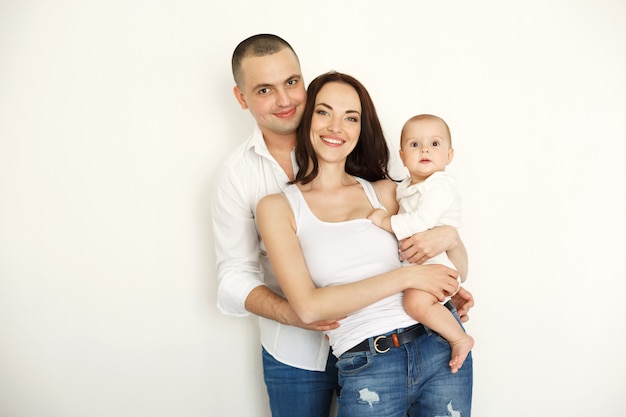 Bella giovane famiglia felice con la posa abbracciante sorridente del piccolo bambino sopra la parete bianca.