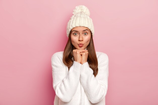 Bella giovane femmina tiene le labbra piegate, le mani sotto il mento, ha un aspetto accattivante nella fotocamera, indossa un abito invernale bianco, ha una pelle sana, una carnagione ben curata, isolato su un muro rosa. Espressioni del viso