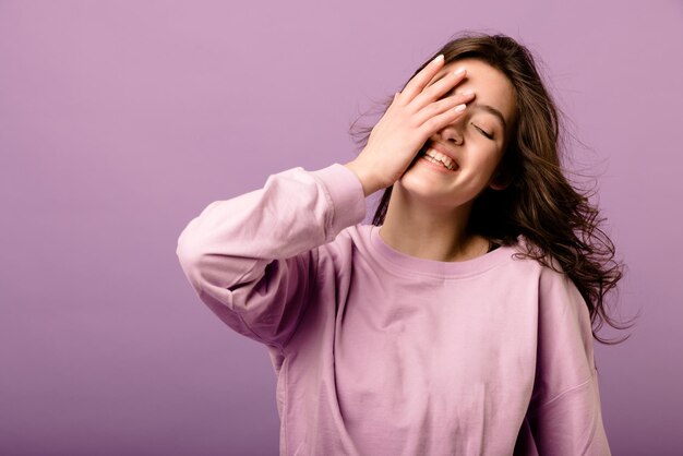 Bella giovane ragazza bruna caucasica sorridente che copre il viso con la mano in piedi su sfondo viola Concetto di buon umore