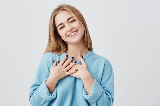 Bella giovane ragazza europea dall'aspetto amichevole positiva con un bel sorriso sincero sentendosi grata e riconoscente, mostrando il suo cuore pieno di amore e gratitudine tenendosi per mano sul seno