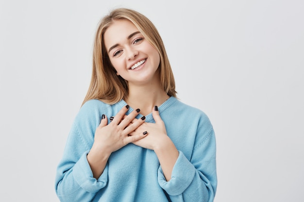 Bella giovane ragazza europea dall'aspetto amichevole positiva con un bel sorriso sincero sentendosi grata e riconoscente, mostrando il suo cuore pieno di amore e gratitudine tenendosi per mano sul seno