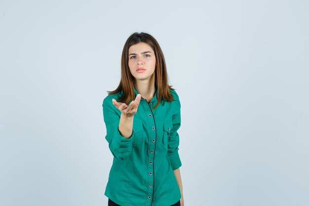 Bella giovane signora che allunga la mano nel gesto interrogativo in camicia verde e che sembra perplessa, vista frontale.