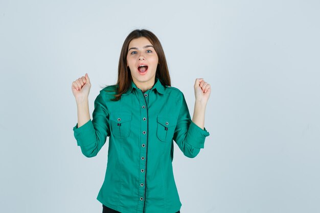 Bella giovane signora in camicia verde che mostra il gesto del vincitore e che sembra felice, vista frontale.