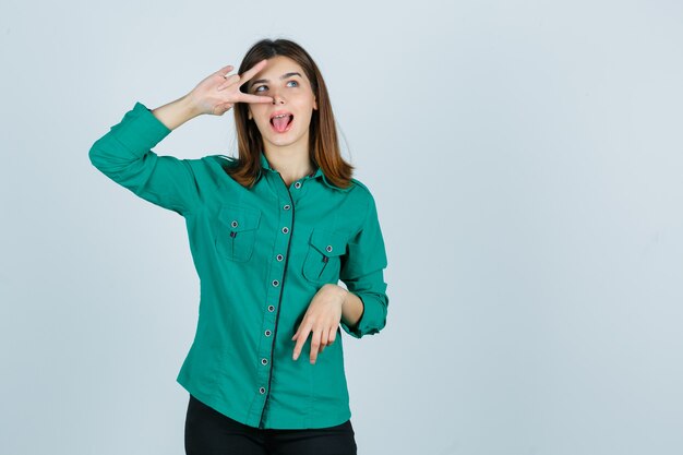 Bella giovane signora in camicia verde che mostra il segno di V sull'occhio e che sembra felice, vista frontale.