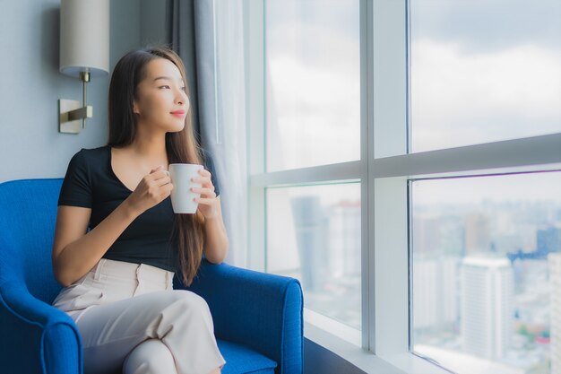 Bella giovane tazza di caffè asiatica della tenuta della donna del ritratto sulla sedia del sofà nell'area del salone