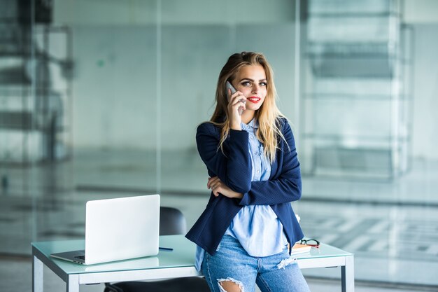 Bella imprenditrice impegnata, in piedi in ufficio, scrivendo nel pianificatore e parlando al telefono cellulare.