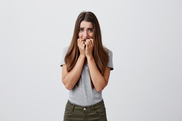 Bella infelice ragazza studentessa caucasica con i capelli lunghi scuri in camicia grigia e jeans rosicchia le dita con espressione spaventata, spaventata da strani suoni