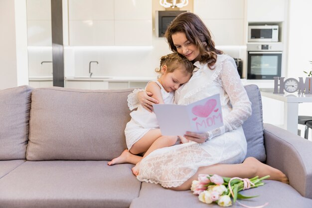 Bella madre e figlia che si siedono sul sofà in salone che legge cartolina d&#39;auguri