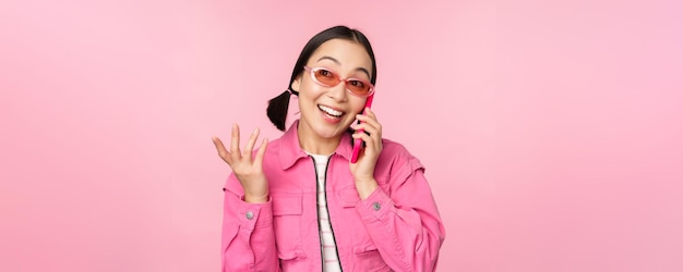 Bella modella coreana in occhiali da sole che parla al telefono cellulare con la faccia felice utilizzando il servizio cellulare per chiamare un amico sullo smartphone in piedi su sfondo rosa